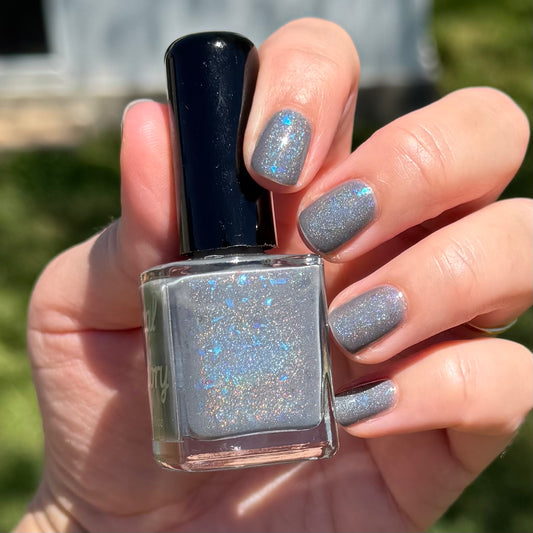 Hand holding a bottle of glittery nail polish with matching nails outdoors. Opal theory polish To Artemis with love