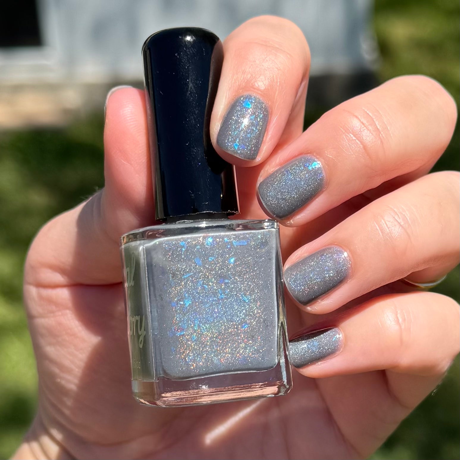 Hand holding a bottle of glittery nail polish with matching nails outdoors. Opal theory polish To Artemis with love