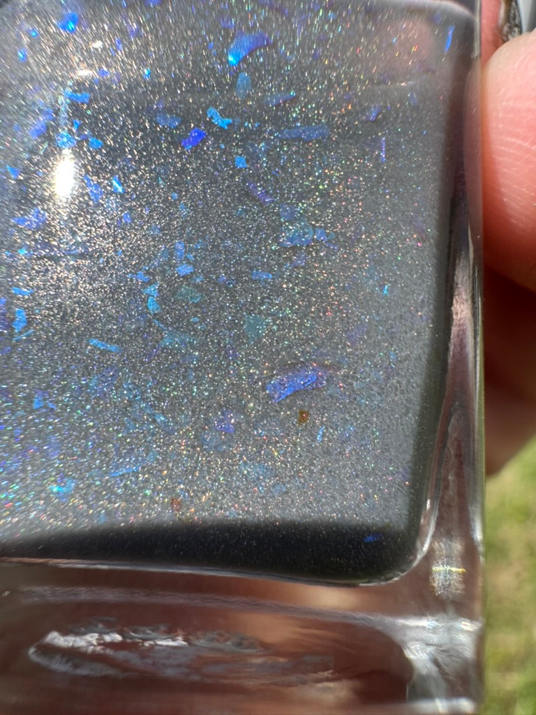 Close-up of a glittery nail polish with blue flakes and silver holographic shimmer. Opal theory polish To Artemis with love