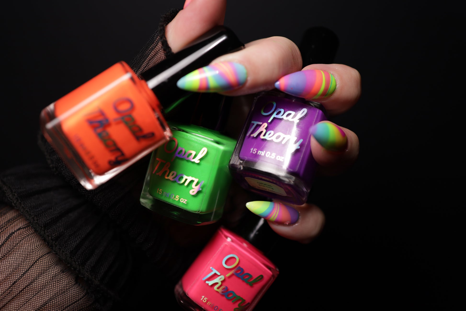 Hand holding four bottles of Opal Theory nail polish with multicolored nails against a black background